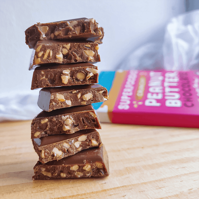 Crunchy Peanut Butter Chocolate Bar 20 x 90g - Superfoodio. Squares of chocolate in a pile, in the background is a full bar of chocolate